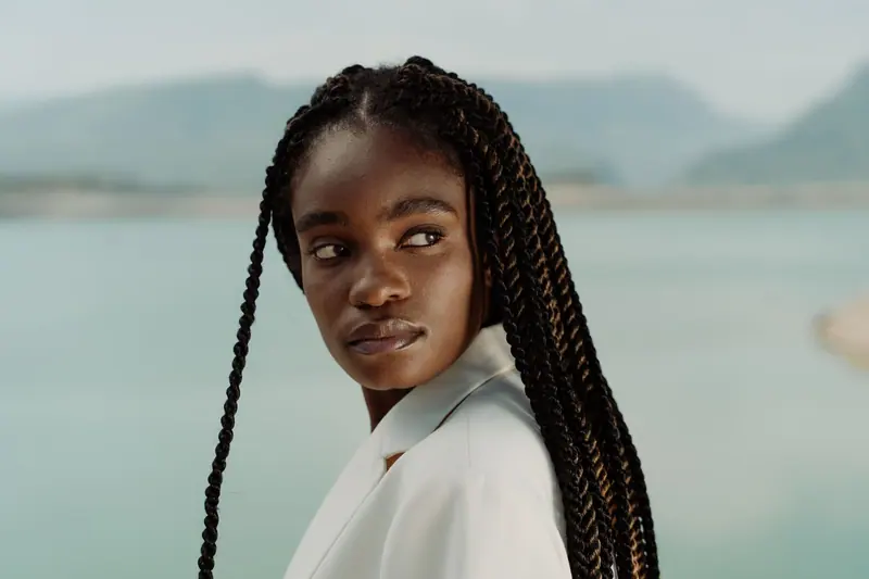 Elegant portrait of a fashionable woman in a white suit with afro braids by a serene water backdrop.