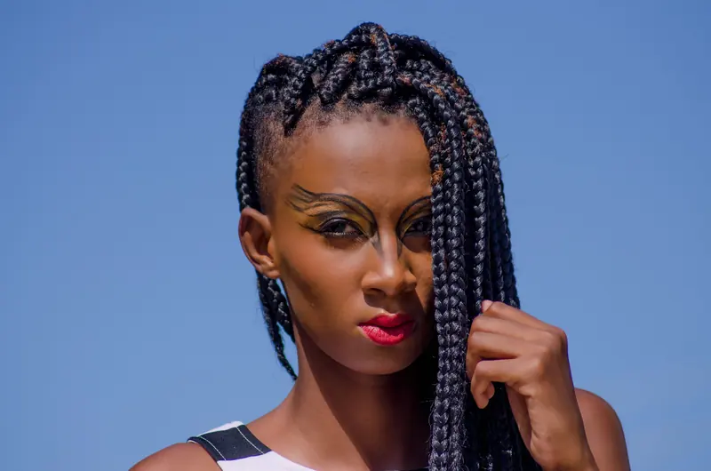 Striking portrait of a woman with bold makeup and intricate braids, set against a clear blue sky.