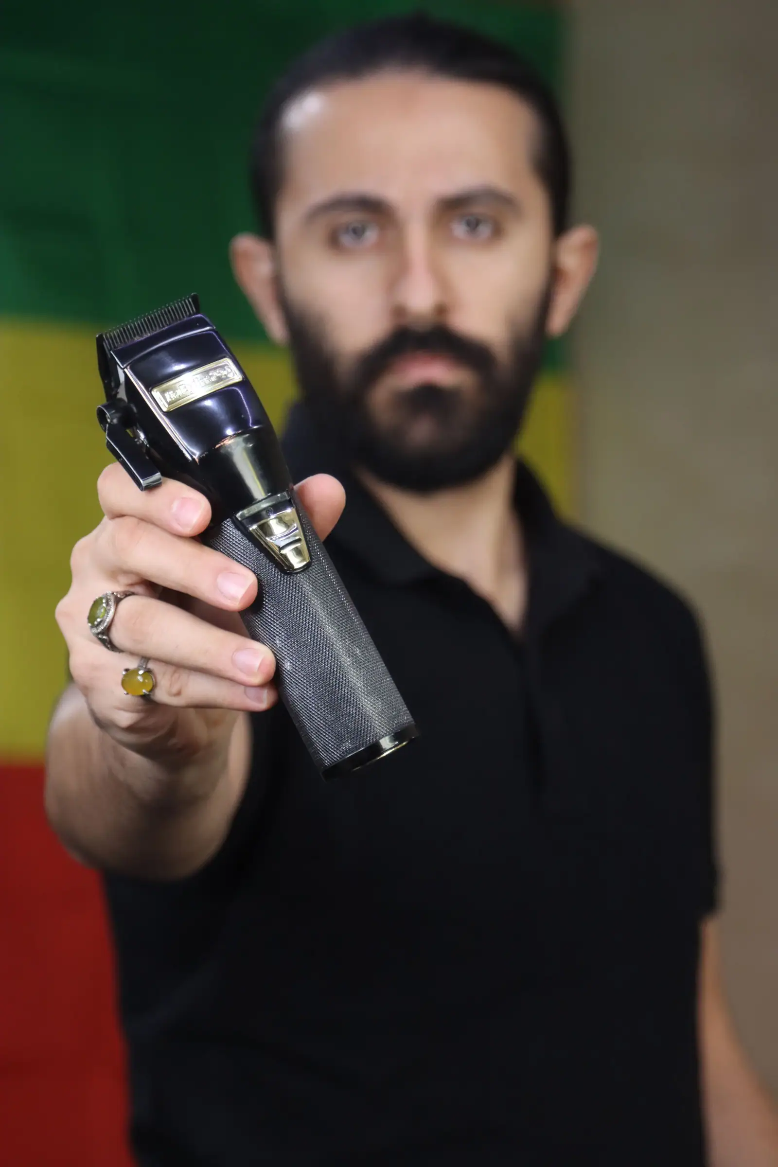 Rami Khoury, a bearded barber, holding his clippers out toward the camera in front of a red, yellow and green wall