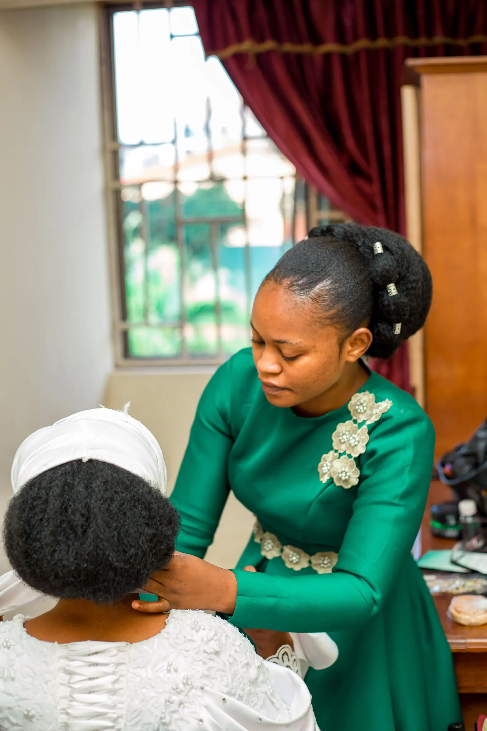 Efua Mensah in a green gown pinning a bride's braided foundation by the window