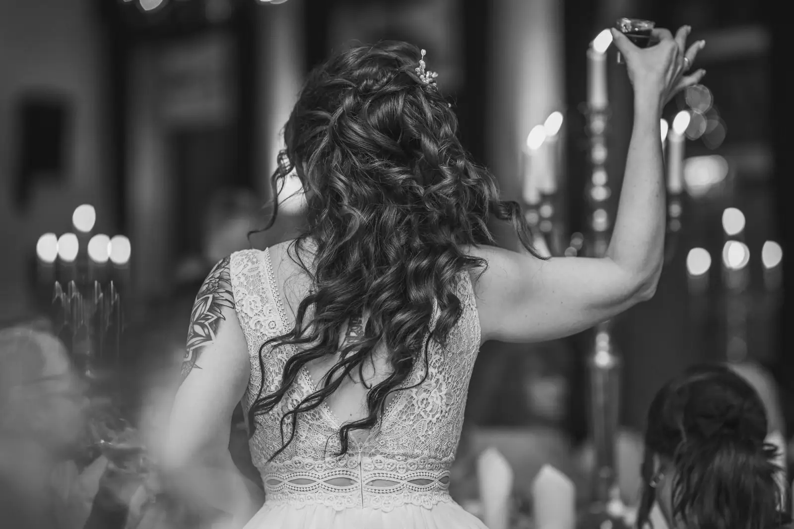 A curly-haired bride from behind raising a toast in a candlelit hall, black and white