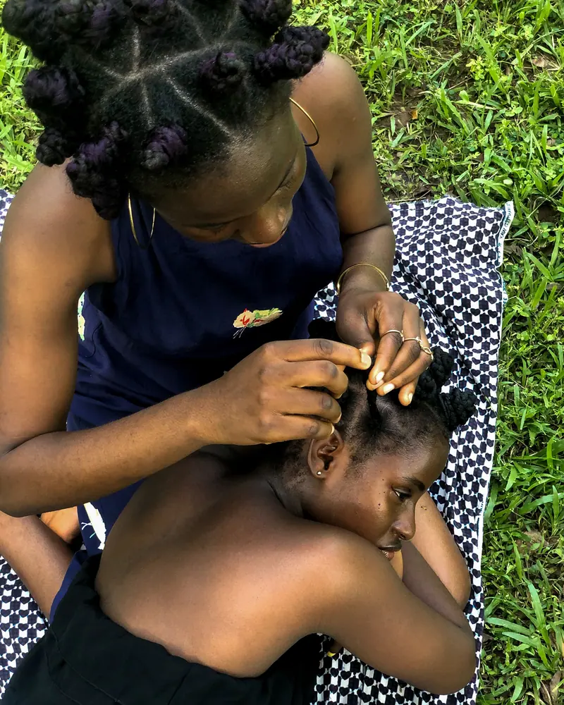 A woman braiding another woman's hair while sitting on a patterned blanket on grass.