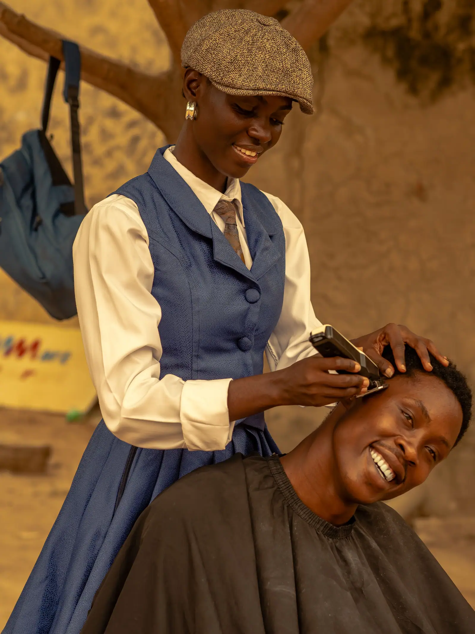Abena Boateng in a tweed cap and vest, laughing with a client while she trims his hair outdoors