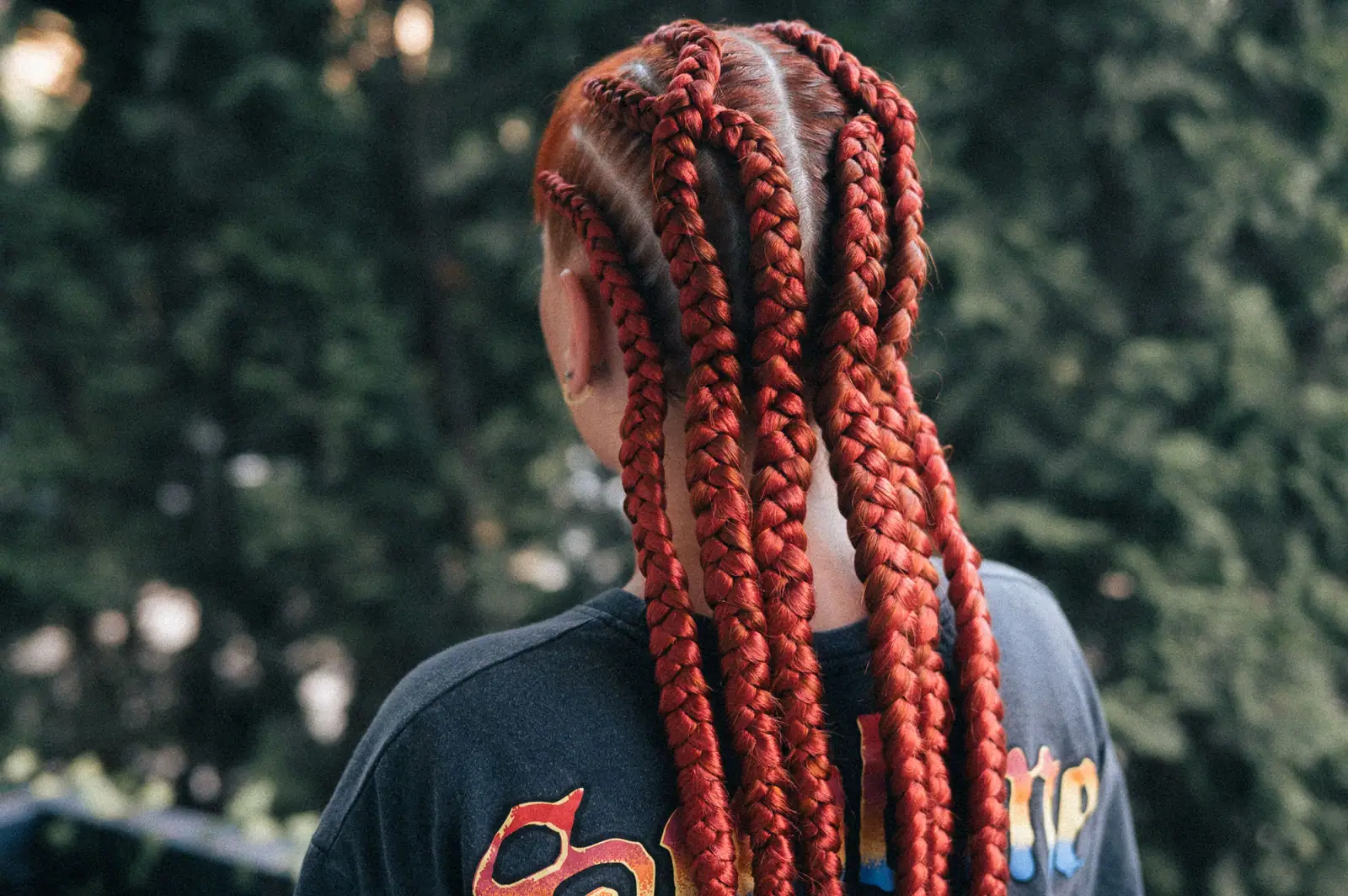 Long red jumbo braids from behind, against dark green leaves