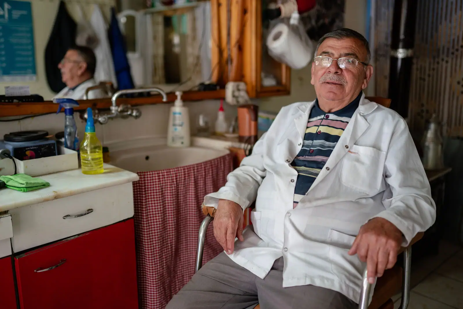 Sal Aydın, an older barber in a white coat, seated in his tiled barbershop