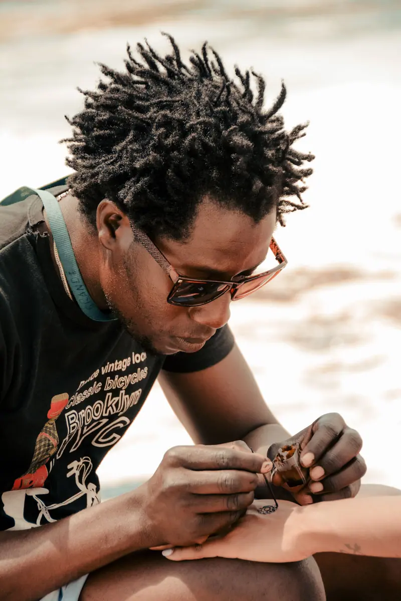 Close-up of an artist applying a detailed henna tattoo on a client's arm. Outdoors setting.
