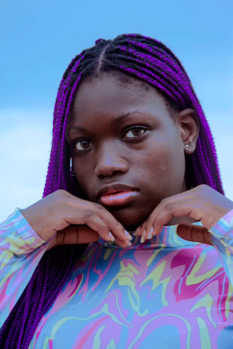 Close-up portrait of a woman with vibrant purple braids posing against a clear blue sky.