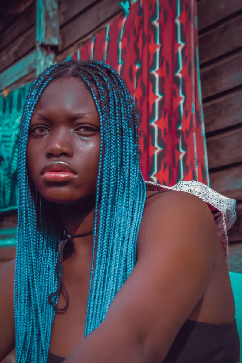 A striking portrait of a woman with blue braids sitting outdoors. Captures a serious expression with vibrant colors.