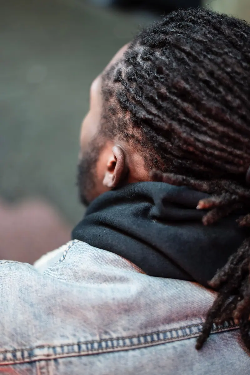A young man with dreadlocks wearing a hoodie and denim jacket from a back view on a city street.