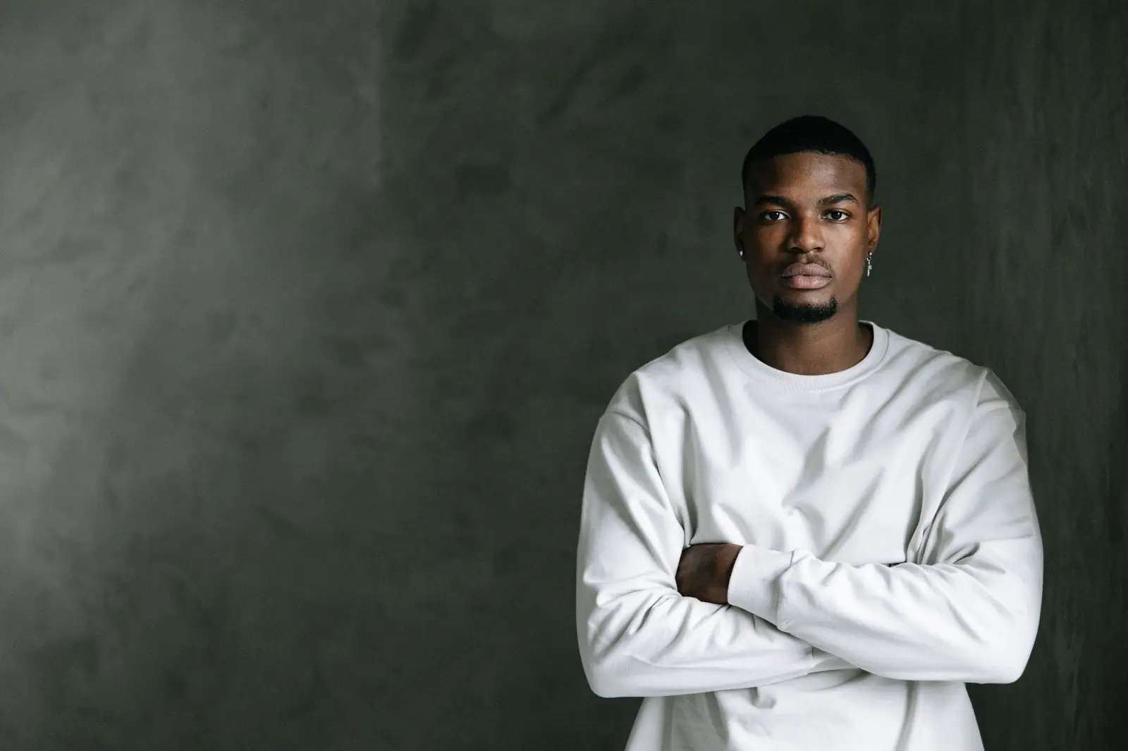 Miles Ashby, a barber in a white sweater, arms crossed against a dark green studio wall