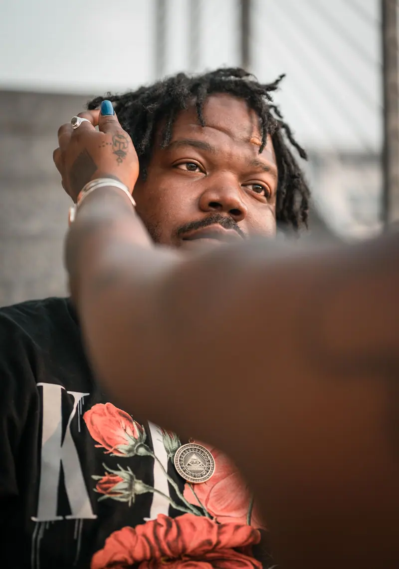 Close-up portrait of a man with dreadlocks outdoors, adorned with a floral shirt and necklace.