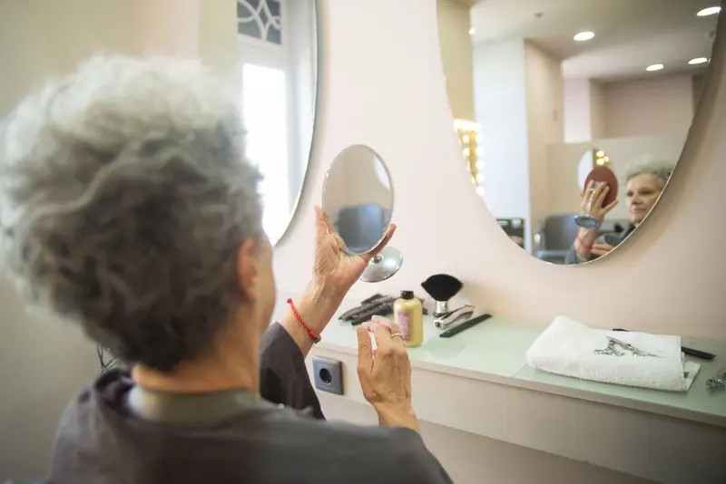 A senior woman inspects her new hairstyle at a salon, reflecting in a handheld mirror.