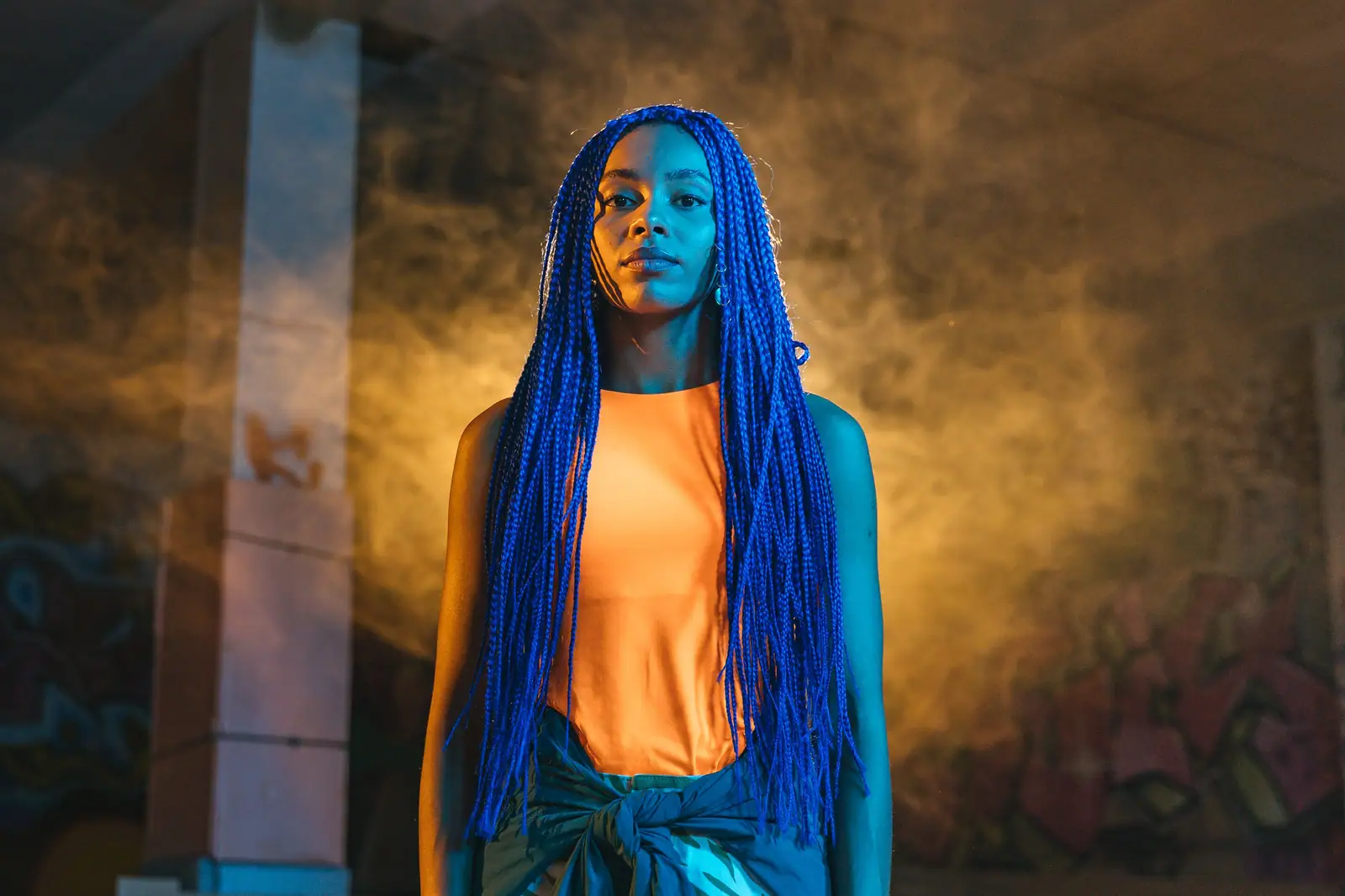 Imani Cole with long cobalt-blue braids and an orange top, in amber smoke at night