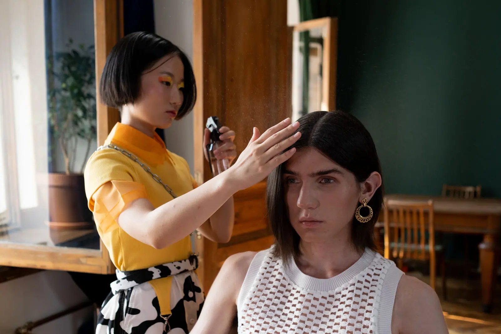 Stylist in a mustard top shaping a client’s dark bob in a green-walled salon