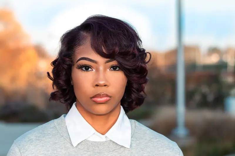 A close-up portrait of a woman with curly hair and a gray sweater, captured outdoors.
