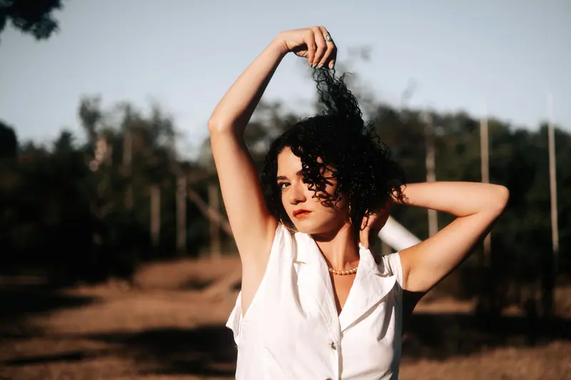 A young woman with curly hair poses outdoors in the countryside during sunset.