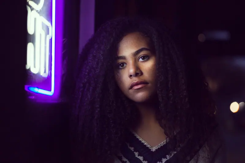 A striking night portrait of a woman with afro hair beside a neon sign.