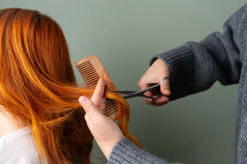 Close-up of red hair being trimmed with scissors and comb on green background.