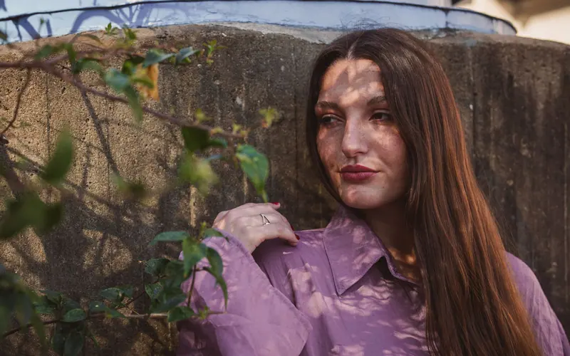Woman outdoors with leaf shadows on face wearing violet shirt, serene expression.
