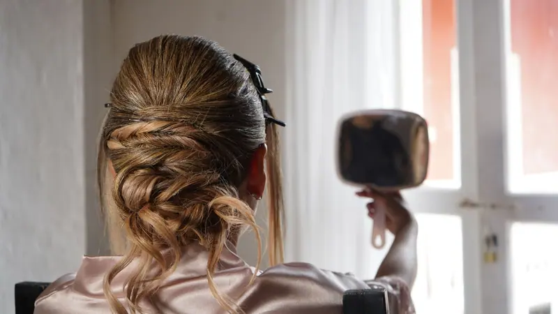 A woman with an elegant braided updo admires her hairstyle in a hand mirror near a window.