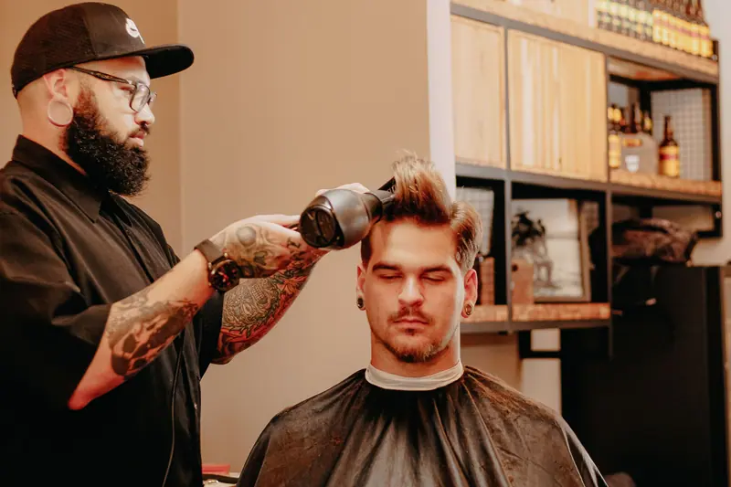 A stylish young man gets his hair dried by a tattooed barber in a modern salon.