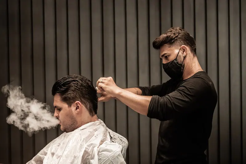A barber wearing a mask trims a customer's hair while the customer exhales smoke indoors.