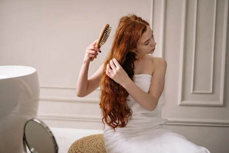 Woman with long red hair wrapped in a towel combing her hair in an elegant bathroom setting.