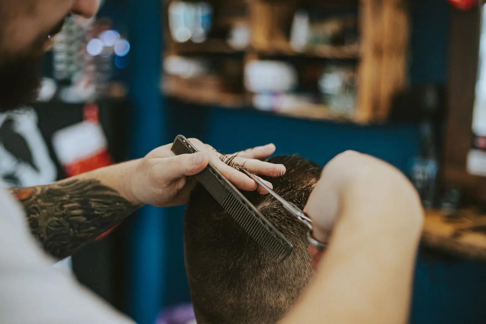 Stylist's tattooed hands cutting a client's hair in a dim, teal-shadowed suite