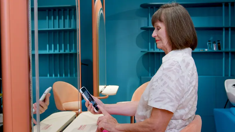 A senior woman sitting in a salon uses her smartphone, reflected in the mirror.
