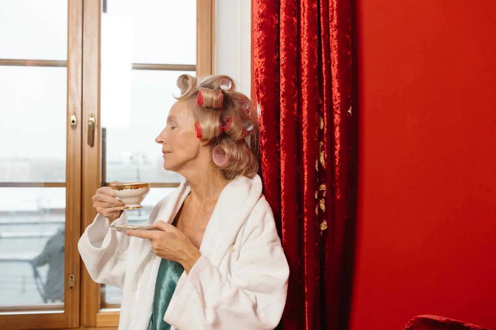 Smiling older woman in a white robe with pink rollers in her hair, sipping tea from a gold-rimmed cup by a window