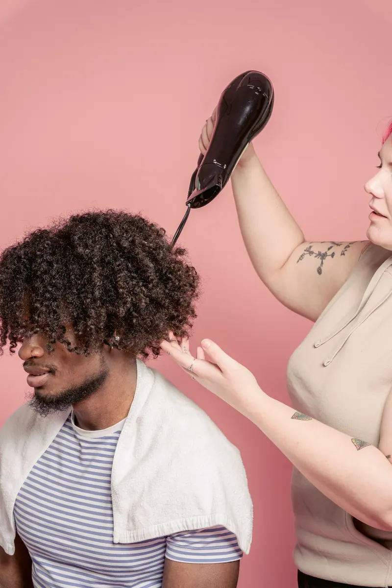 Stylist working on a man's curly hair in a studio with a pink background.