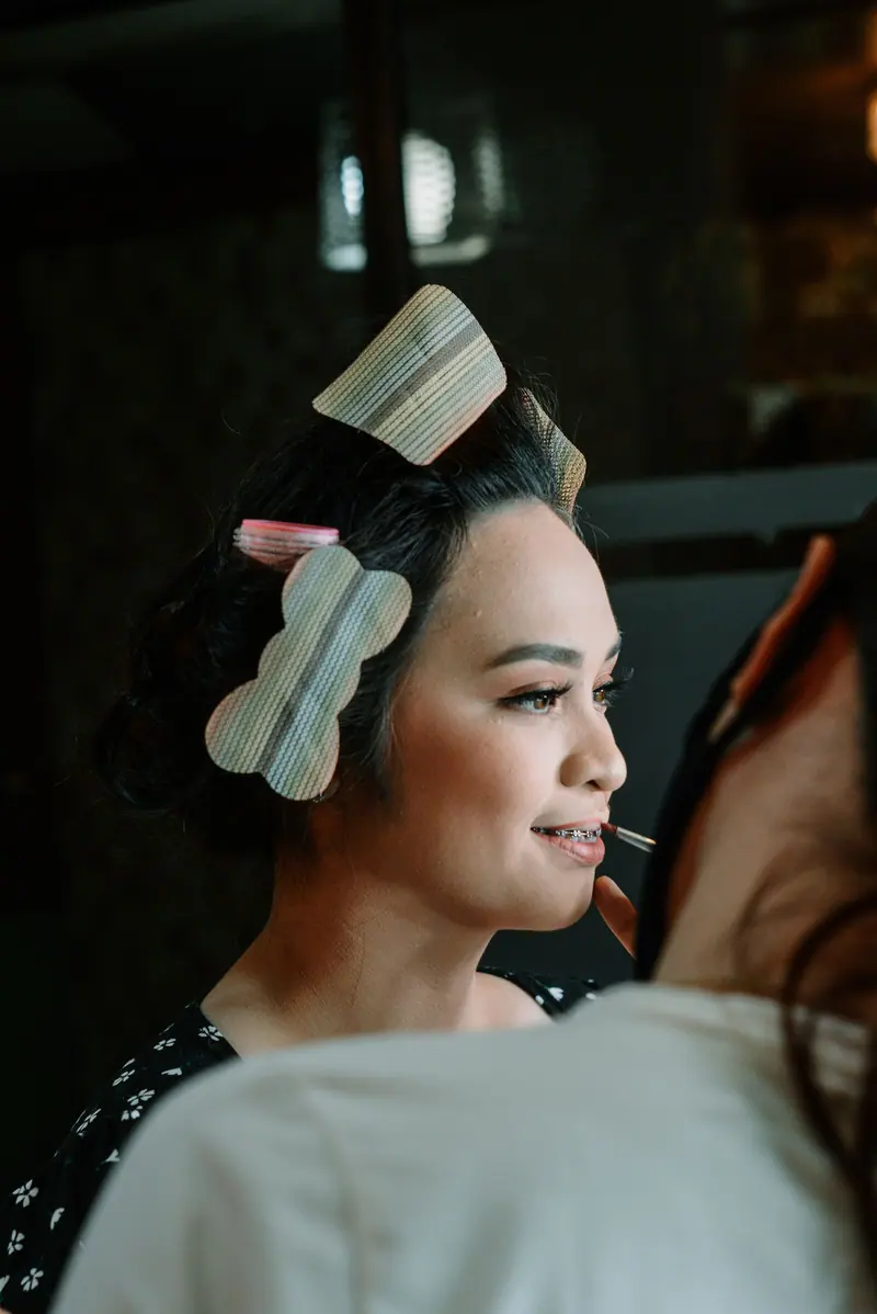 A woman getting makeup applied with hair rollers in a beauty salon setting.