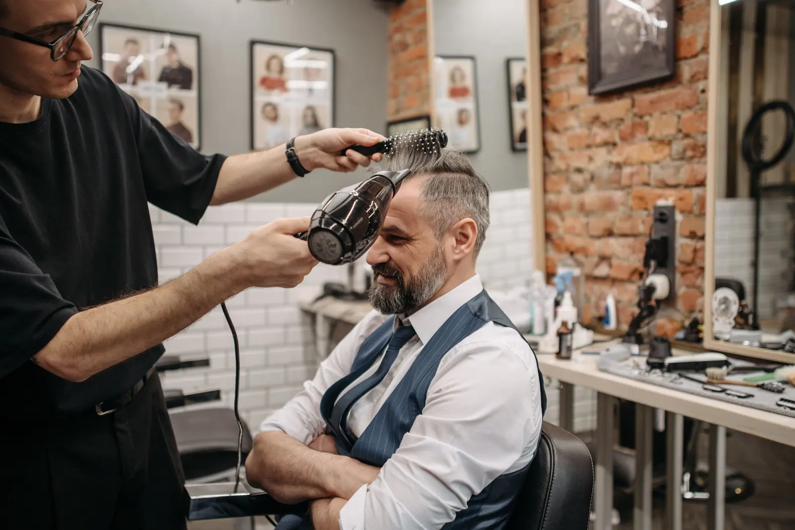 Barber blow-drying a seated client's hair in a brick-walled grooming suite
