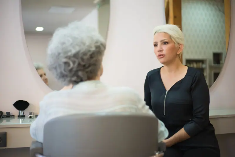 Two women having a conversation in a salon setting with a mirror reflecting their interaction.