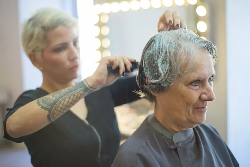 A hair stylist trims the hair of a smiling senior woman in a salon.