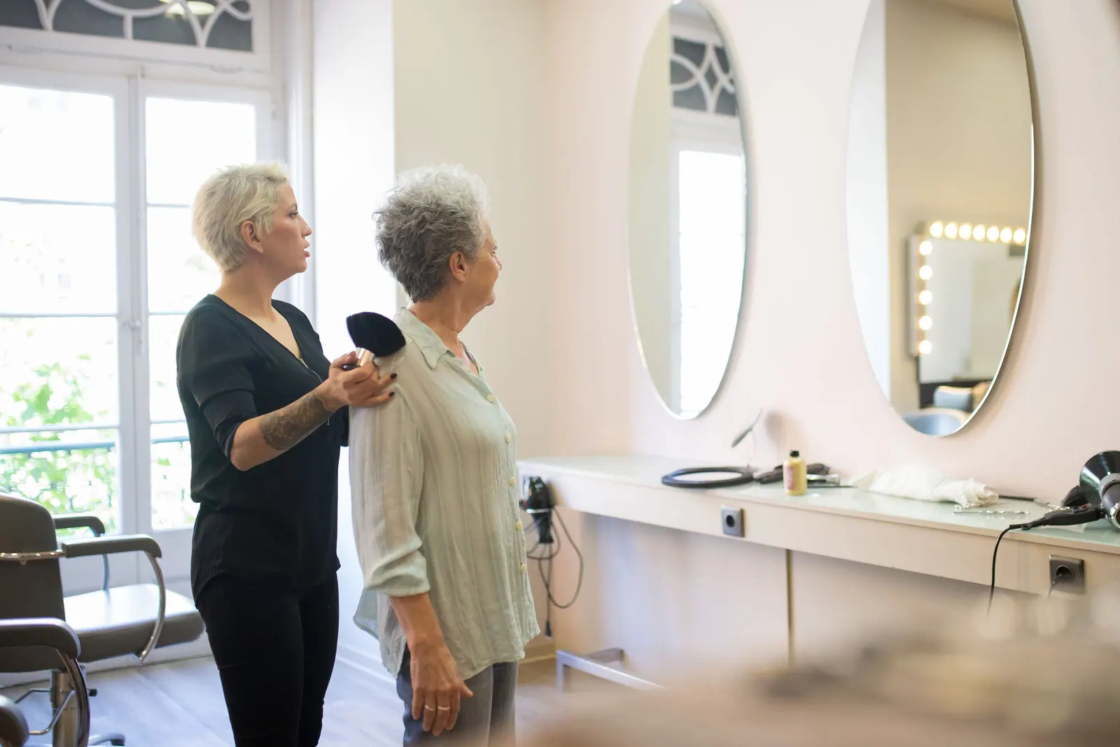 Stylist with a senior client in an airy white salon suite with oval mirrors