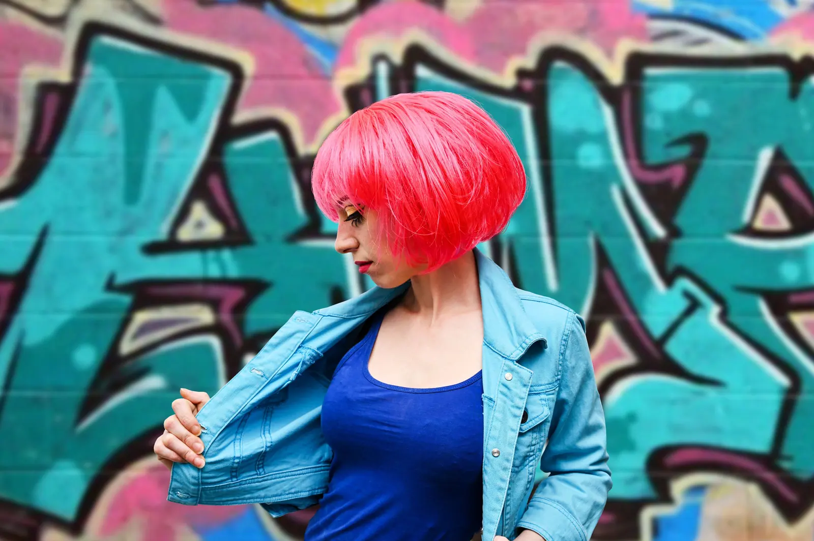Woman with a glossy coral-pink bob and a denim jacket in front of a teal and pink graffiti wall
