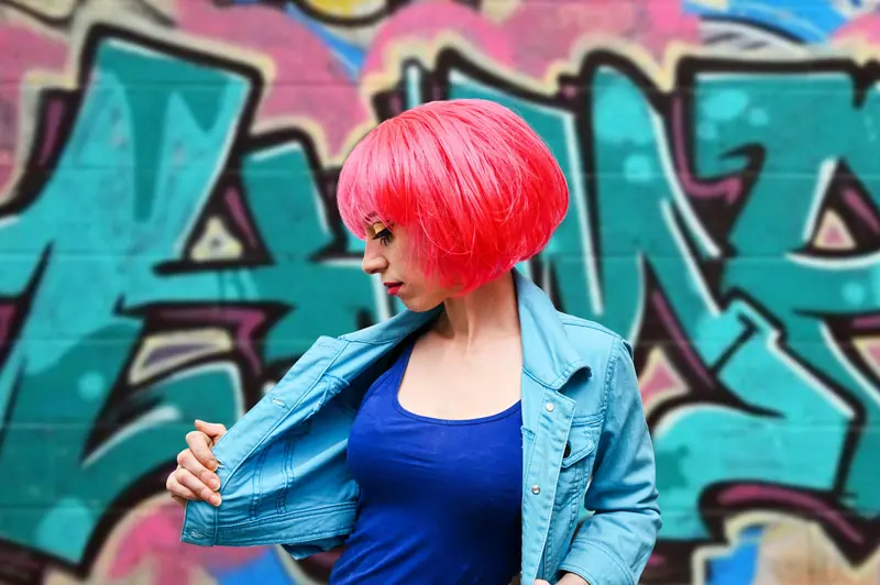 Stylish woman with pink hair poses in front of colorful graffiti mural.