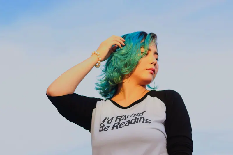 A young woman with teal hair wearing a reading-themed shirt poses confidently outdoors.