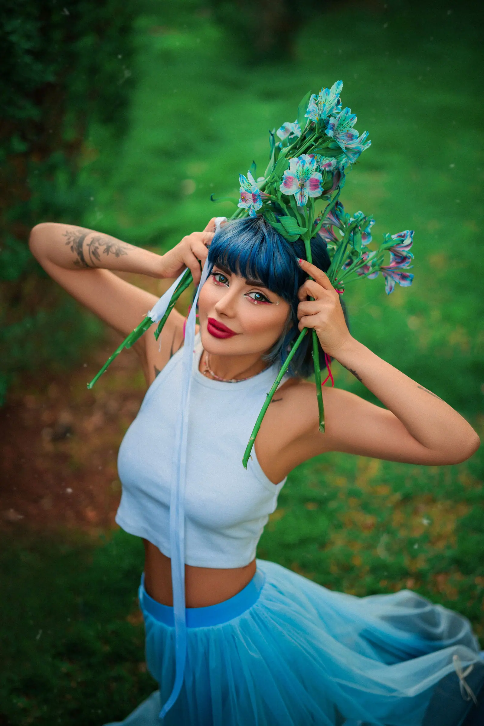 Colorful fashion portrait of a woman with dyed blue hair and floral headpiece in a garden setting.