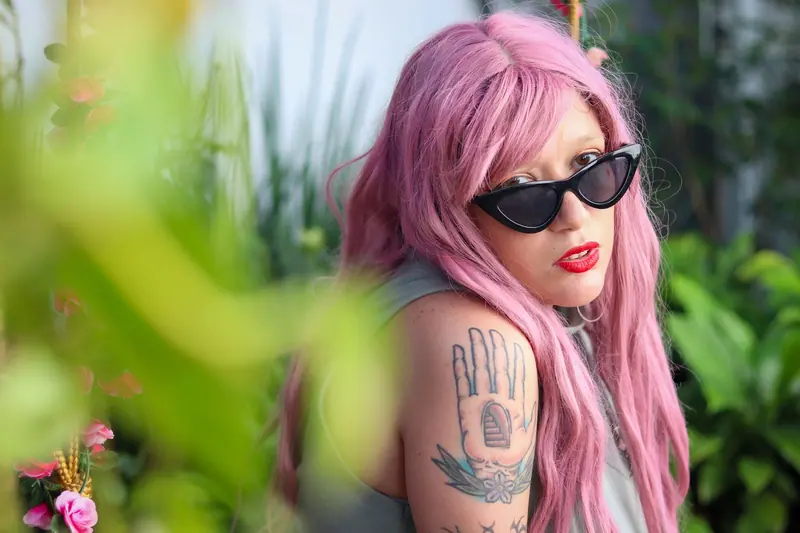 Stylish woman with pink hair and tattoos wearing sunglasses in a lush garden setting.