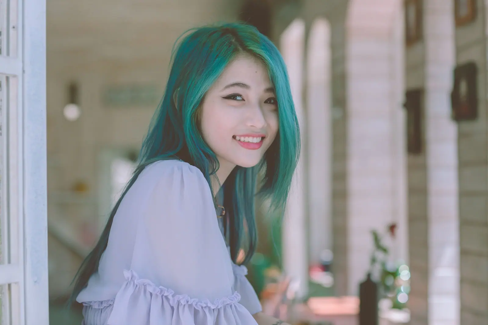Smiling woman with long verdigris-teal hair in a lilac blouse beside a sunlit window