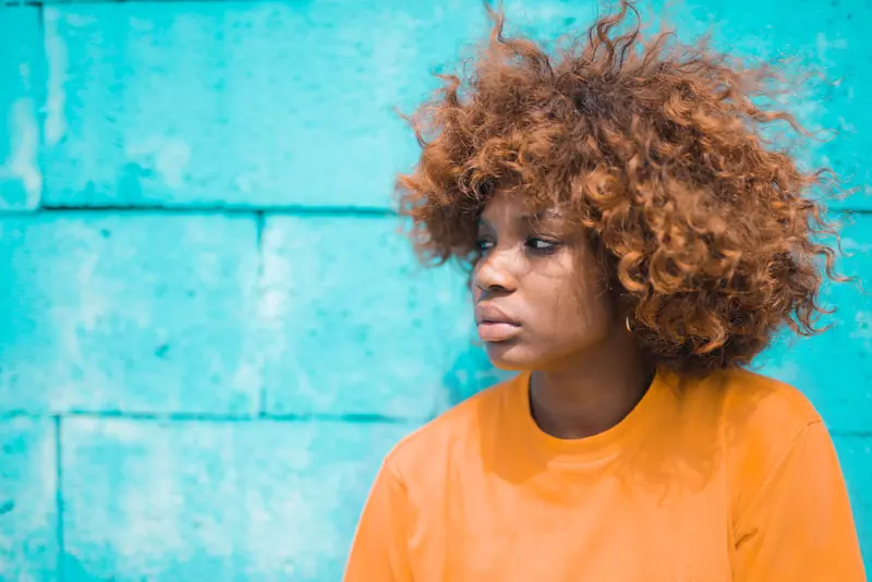 A young woman with natural curly hair stands against a bright blue wall, wearing a casual orange top.