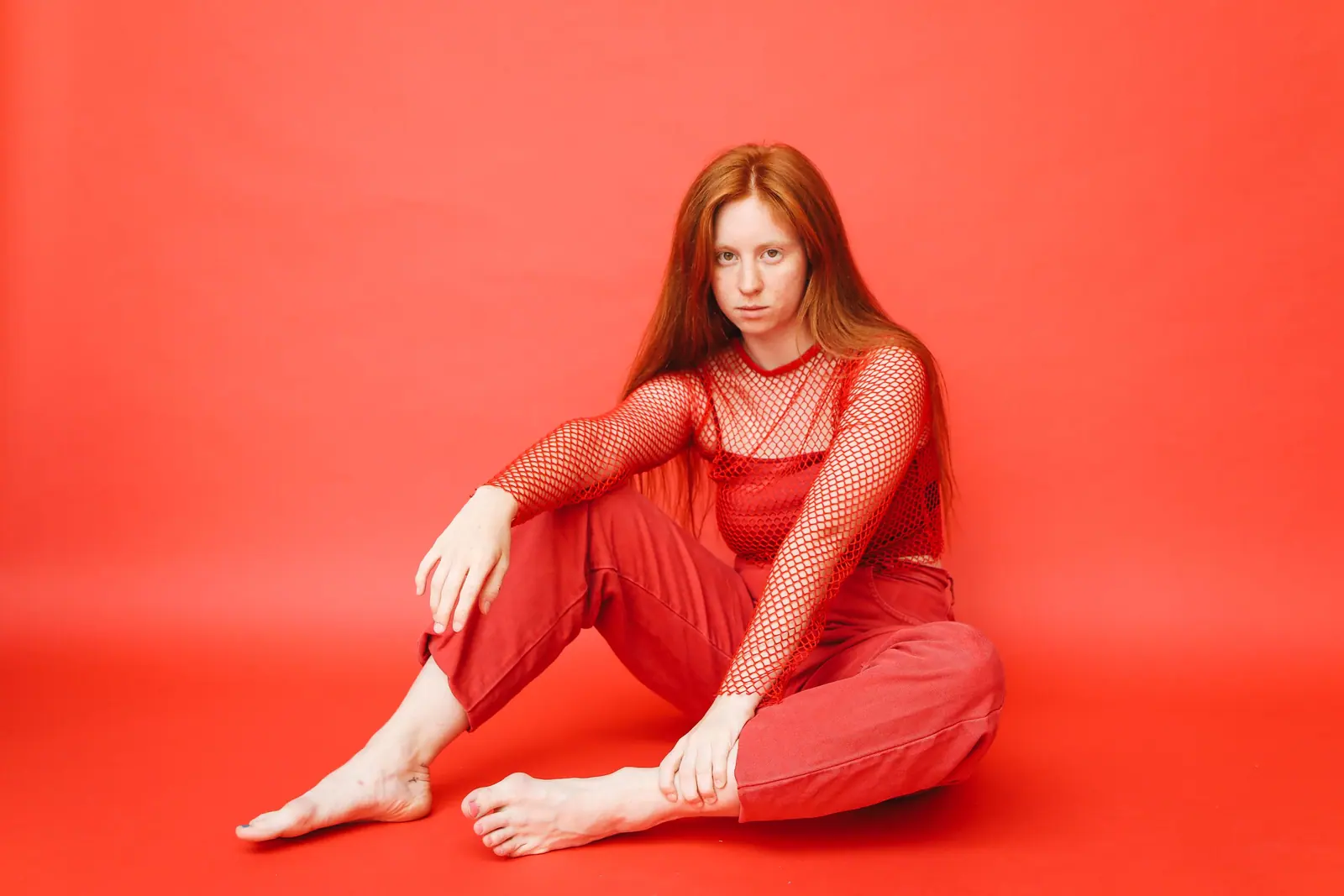 Woman with long copper-red hair in a red mesh top sitting barefoot against a bright red backdrop