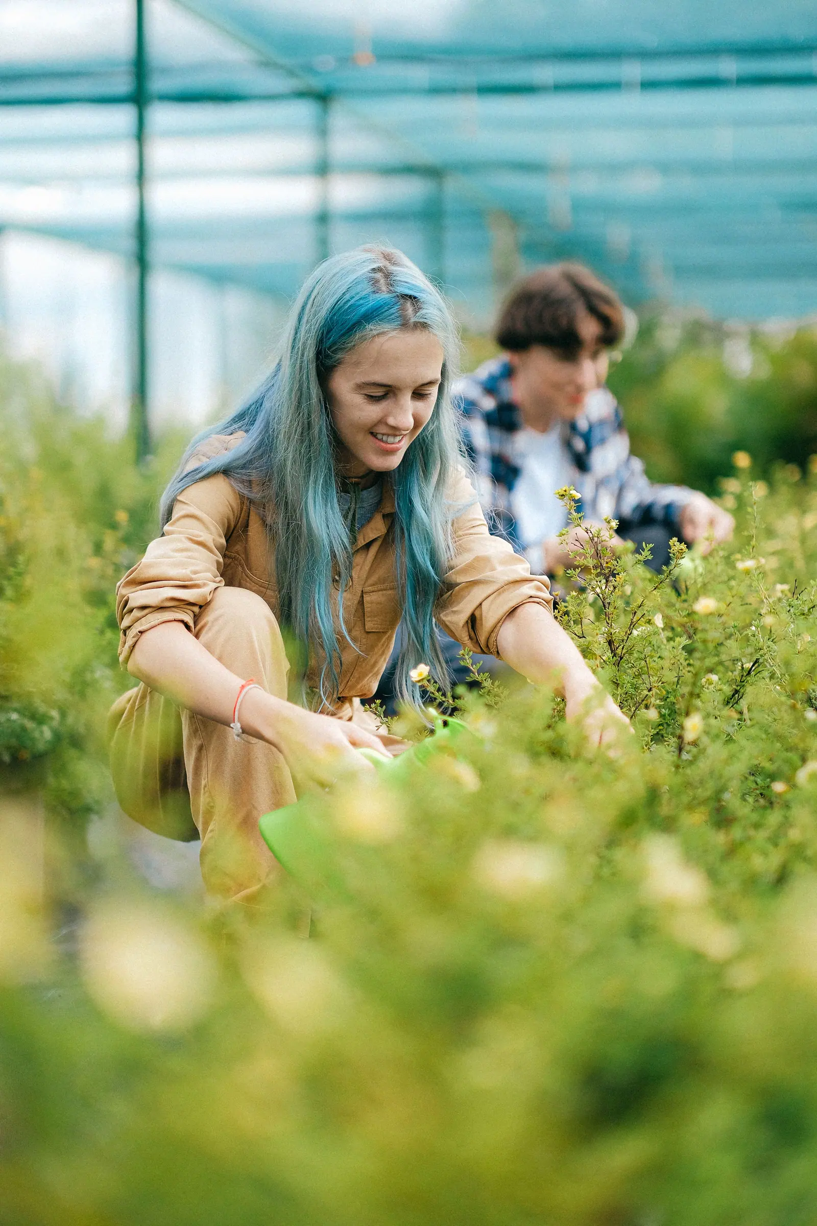 Adult with blue hair gardening in a greenhouse, surrounded by greenery and flowers, enjoying nature.