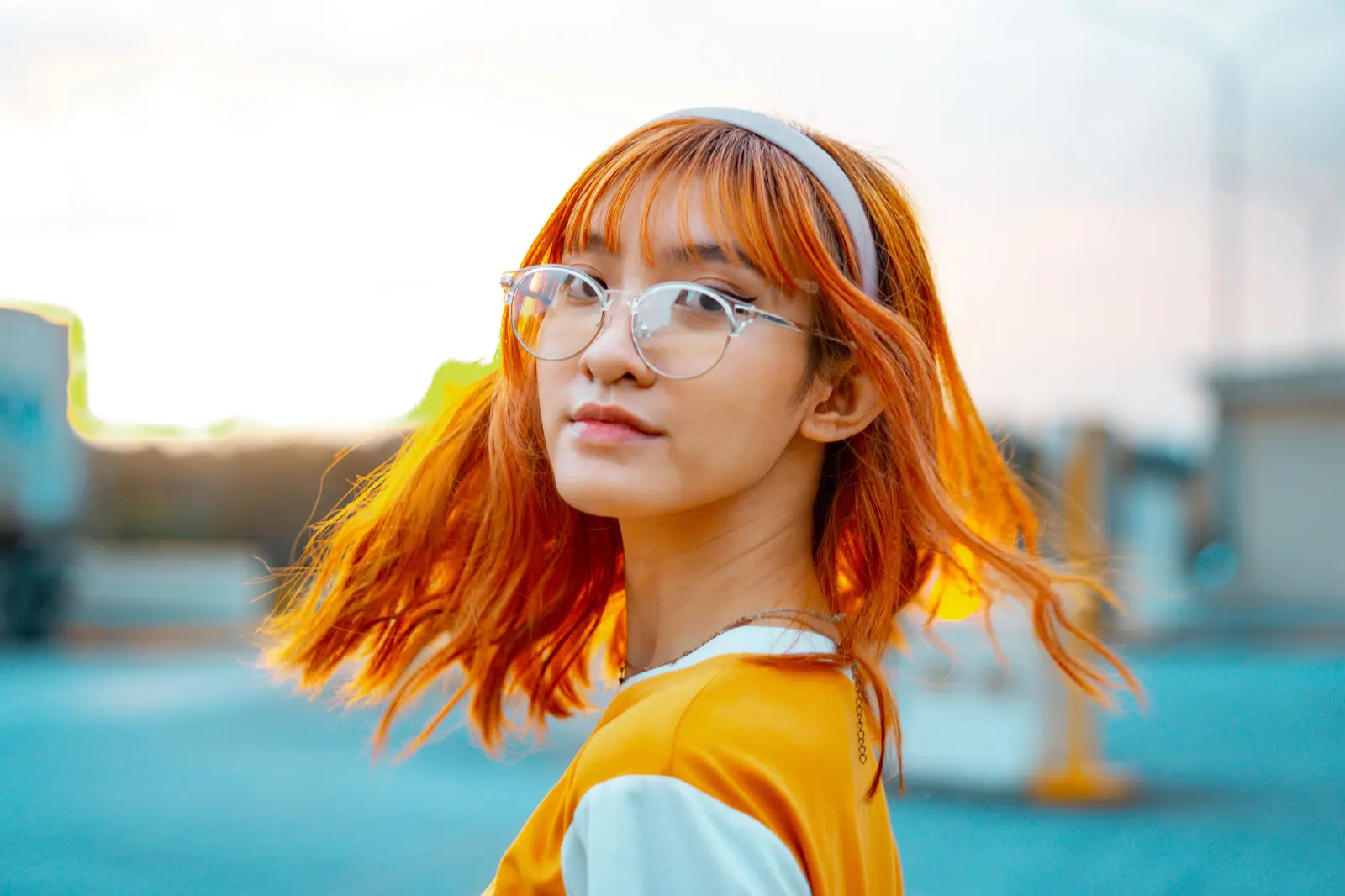 Woman with bright tangerine hair and a white headband turning over her shoulder against a cyan sky