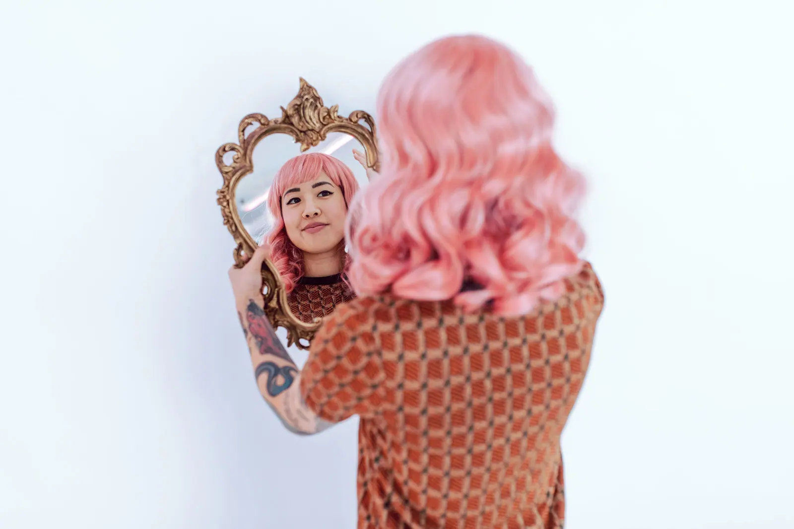 Asian woman with pink hair and tattoos admires reflection in ornate mirror on white background.