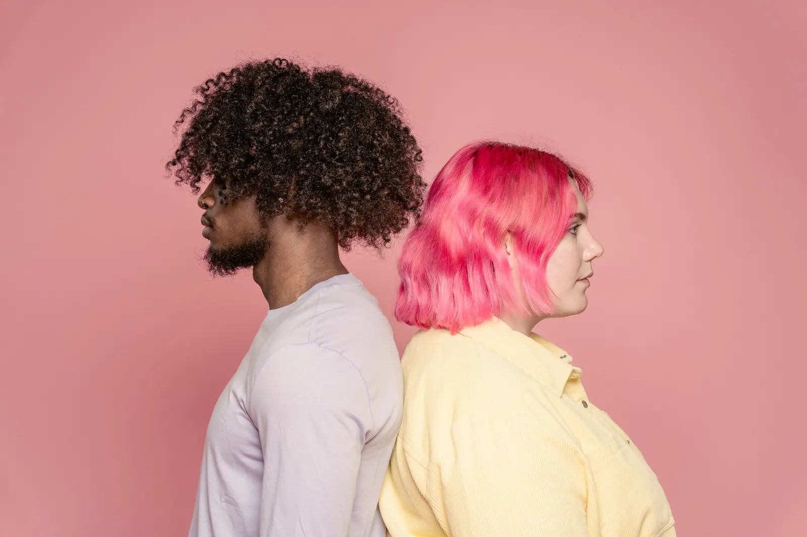 Artistic portrait of a diverse couple back to back against a pink background, showcasing unique hairstyles and fashion.