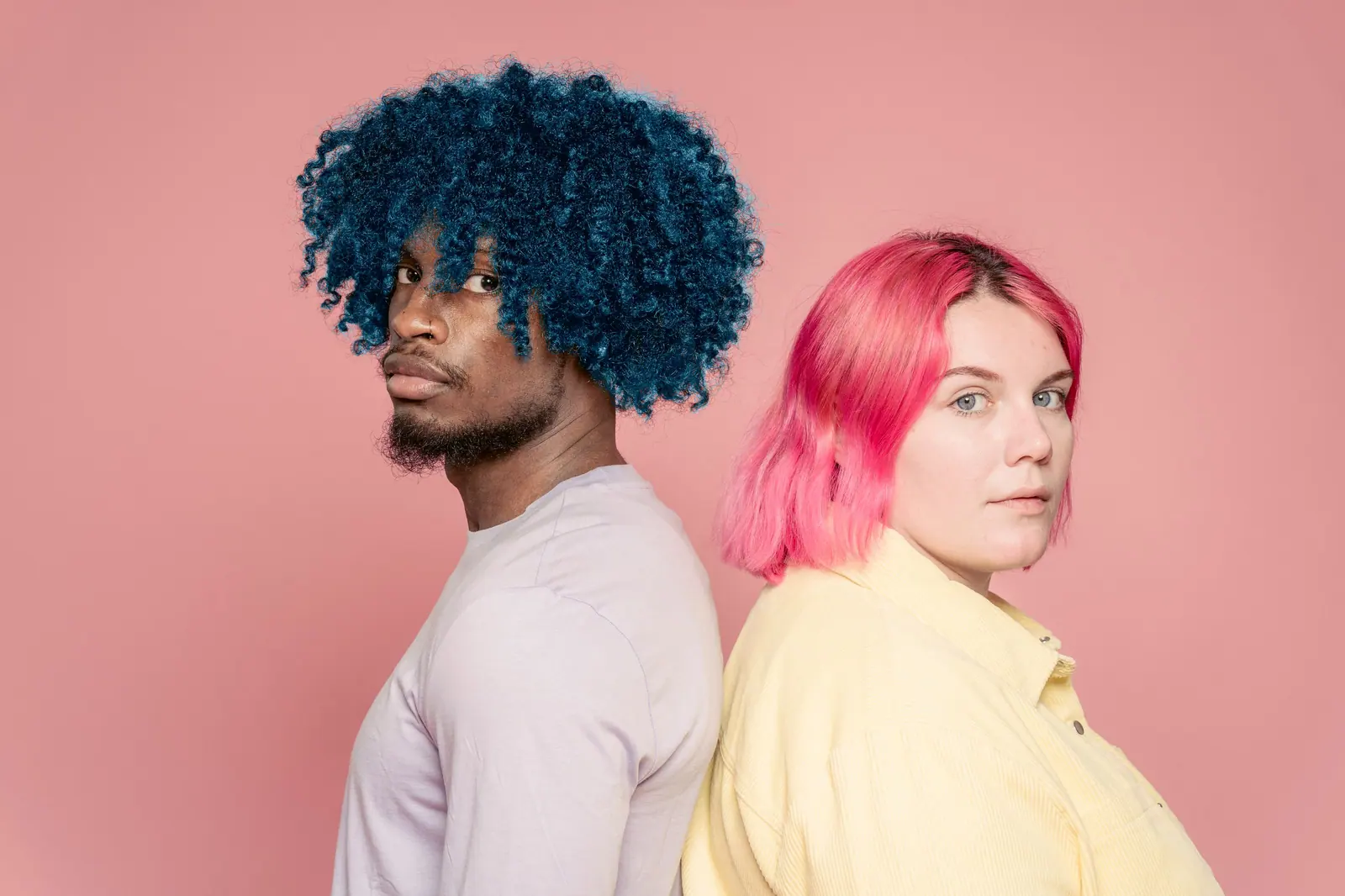 Two models back to back, one with a deep-blue afro and one with a glossy pink bob, against a dusty-pink wall