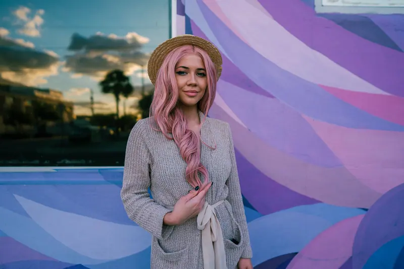 Stylish woman with pink hair poses in front of a colorful mural during the day.